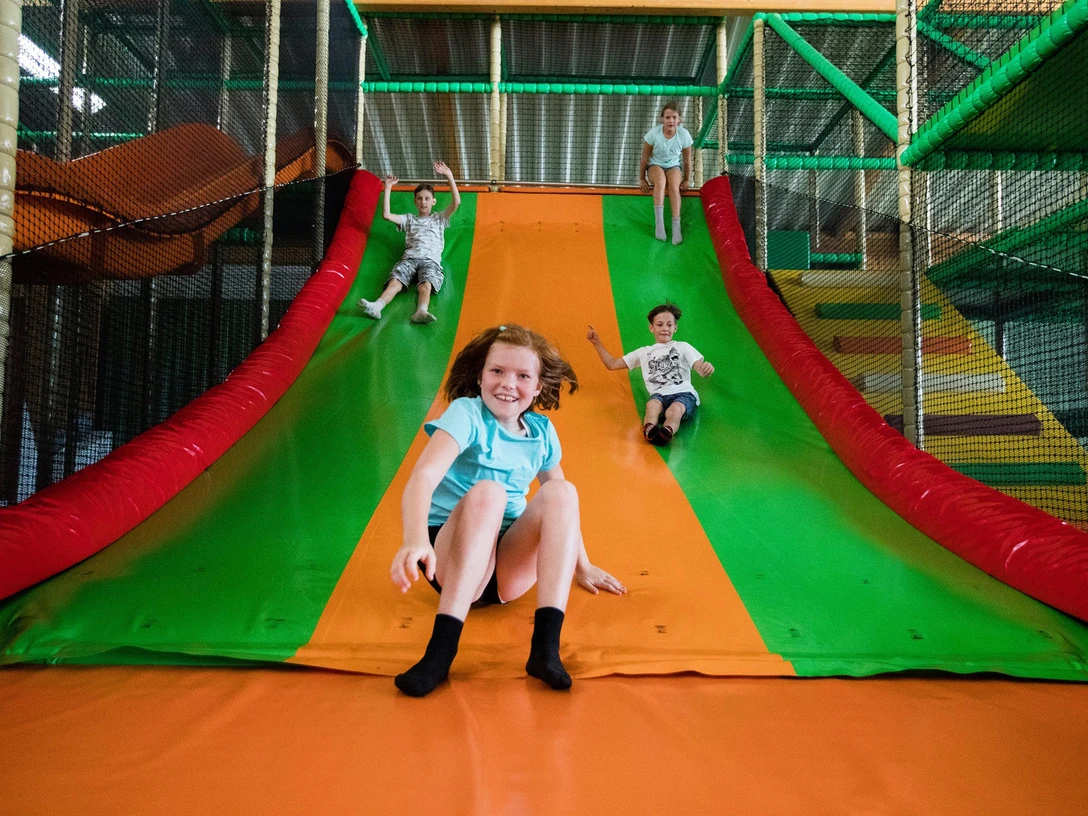Rutsche im Solivital Sebnitz Vier Kinder rutschen lachend eine große, bunte Rutsche in einem Indoor-Spielplatz hinunter. Die Farben der Rutsche sind grün und orange.