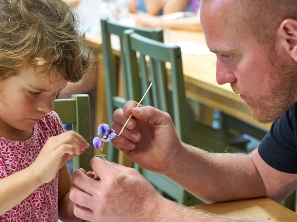 A child and an adult look intently at a small, purple flower; the adult holds a paintbrush, the atmosphere is calm and instructive.