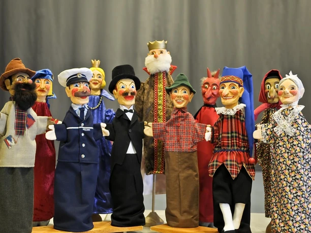 Loutky Punch and Judy z divadla Maxe Jacoba Skupina barevných ručních loutek, včetně policisty, krále a čerta, na šedém pozadí. Loutky mají veselé výrazy ve tváři.
