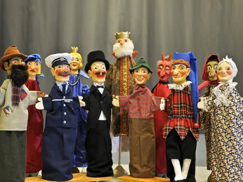 Punch and Judy Puppets from the Max Jacob Theatre Eine Gruppe bunter Handpuppen, darunter ein Polizist, ein König und ein Teufel, vor einem grauen Hintergrund. Die Puppen tragen fröhliche Gesichtsausdrücke.