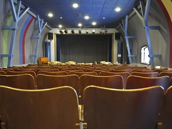 Saal im Max Jacob Theater Leerer Theatersaal mit braunen Holzstühlen, einer Bühne mit geschlossenem Vorhang und bunten Streifen an den Wänden, ruhige Atmosphäre.
