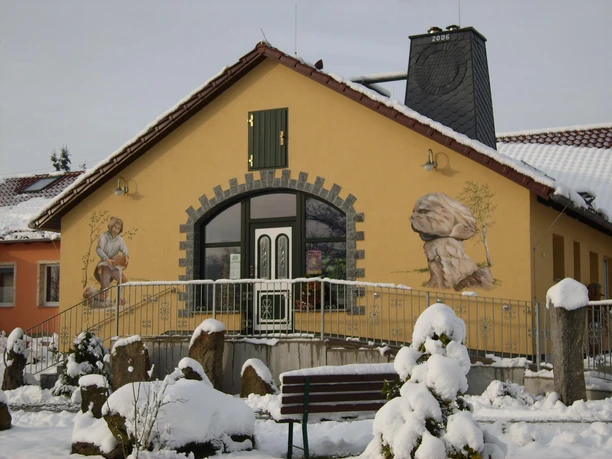 Yellow building with painted facade, showing a person and rocks, surrounded by snow-covered bushes and stones, wintry atmosphere.