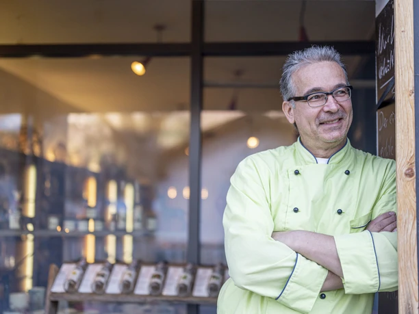 Marys Marmeladen Ein Mann im grünen Kochmantel steht vor einem Geschäft und lächelt. Im Hintergrund sind Regale.