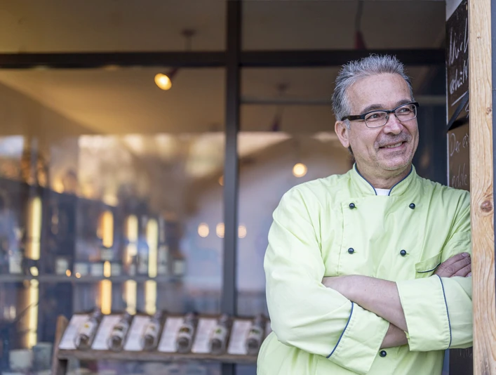 Marys Marmeladen Ein Mann im grünen Kochmantel steht vor einem Geschäft und lächelt. Im Hintergrund sind Regale.