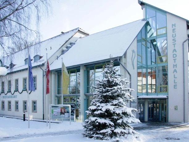 Zasněžená Neustadthalle s prosklenou fasádou, lemovanou vlajkami a zasněženým vánočním stromem, pod jasnou oblohou.