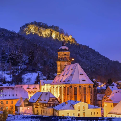 Königstein im Winter Verschneite Stadtansicht bei Dämmerung mit beleuchteter Kirche im Vordergrund und erleuchtetem Schloss auf einem Hügel im Hintergrund.