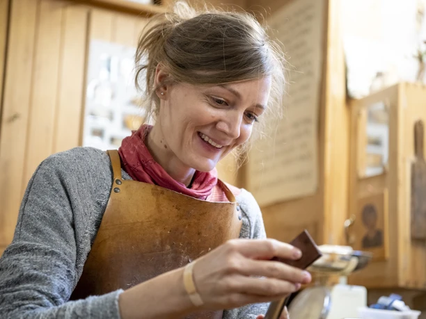 Holzdesign & Holzobjekte Eva Engler Inser Hoamat Partner Eine Frau arbeitet in einer Holzwerkstatt, trägt eine Lederschürze und schaut auf ein Werkstück.