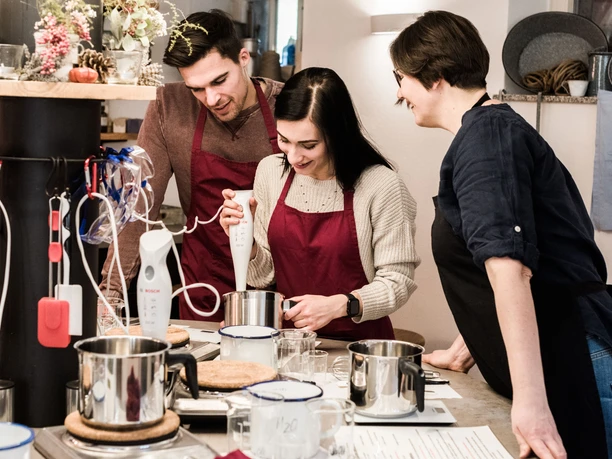 Three people in red aprons cooking together in a kitchen, using a hand blender and looking concentrated and cheerful.