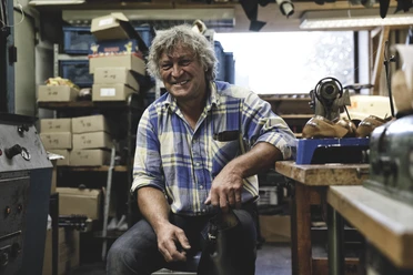 Zollner Schuhe Inser Hoamat Partner Handwerker in einer gut ausgestatteten Werkstatt lächelt, umgeben von Werkzeugen und Schuhmaterialien.