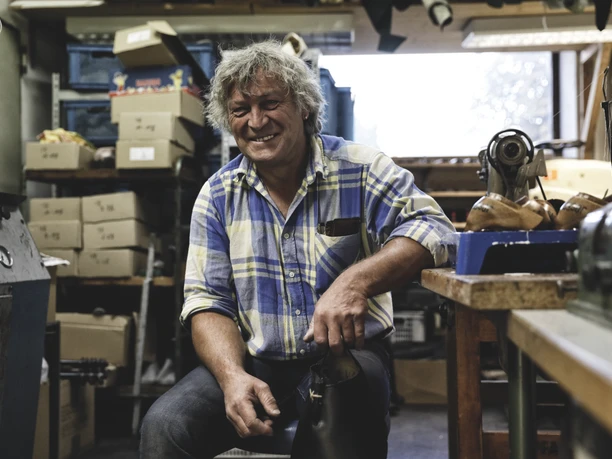 Zollner Schuhe Inser Hoamat Partner Handwerker in einer gut ausgestatteten Werkstatt lächelt, umgeben von Werkzeugen und Schuhmaterialien.