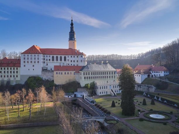 Historický hrad Weesenstein s červenou střechou a vysokou věží, obklopený upravenými zahradami a holými stromy pod jasně modrou oblohou.