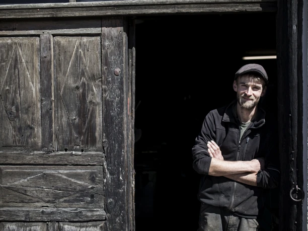Aberl Metallgestaltung Ein Mann lehnt lächelnd in einer Balkontür eines alten Holzgebäudes.