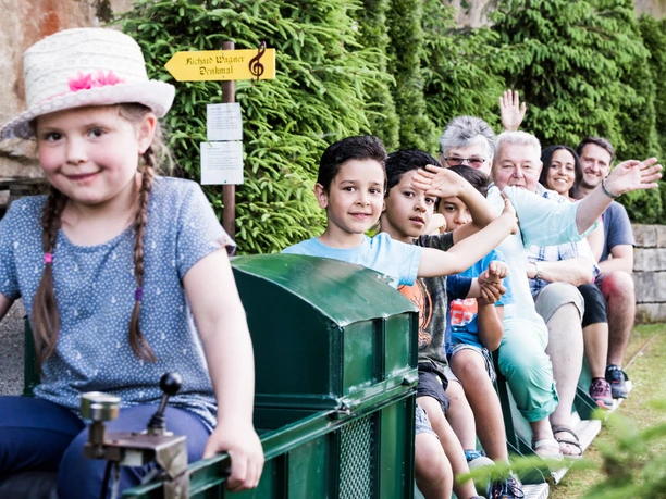 kleine Eisenbahn im Miniaturpark Eine Gruppe von Menschen, darunter Kinder und Erwachsene, sitzt fröhlich winkend auf einem kleinen Zug; im Hintergrund sind grüne Büsche und ein Schild zu sehen.