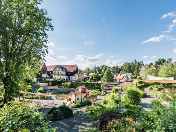 Zelený park s dětským hřištěm, obklopený hrázděnými domy a stromy, pod jasnou modrou oblohou; uvolněná, idylická atmosféra.