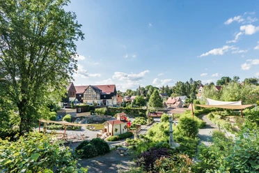 Miniaturpark kleine Sächsische Schweiz Grüner Park mit Spielplatz, umgeben von Fachwerkhäusern und Bäumen, unter klarem, blauem Himmel; entspannte, idyllische Atmosphäre.