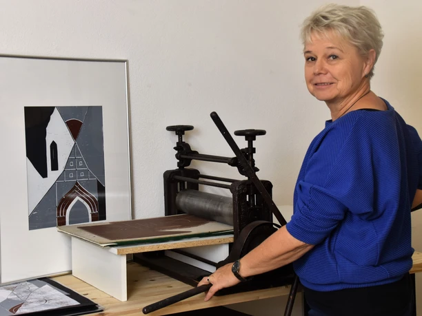 Een vrouw in een blauwe trui staat bij een drukmachine, naast haar een ingelijst abstract kunstwerk in grijs- en bruintinten.