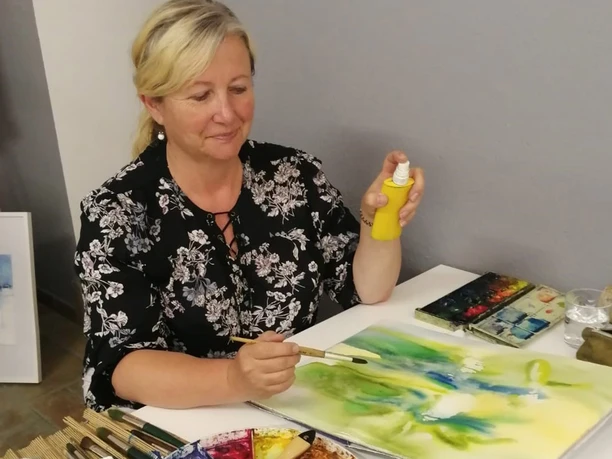 Een vrouw in een zwarte blouse schildert met aquarelverf op papier en houdt een gele spuitfles vast; kwasten en verf liggen verspreid op tafel.