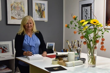 Vrouw met blond haar in een blauw topje staat aan een tafel met tekenmateriaal, omringd door kunstwerken en een boeket bloemen in een vaas.