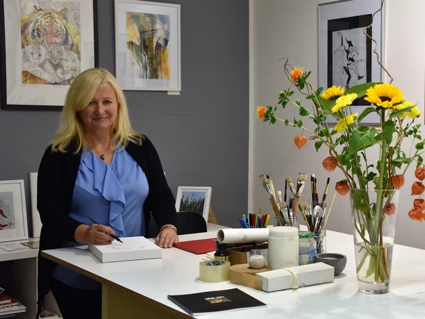 Vrouw met blond haar in een blauw topje staat aan een tafel met tekenmateriaal, omringd door kunstwerken en een boeket bloemen in een vaas.