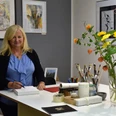Aquarellkurs Galeriewerkstatt Ansichtssache Frau mit blonder Haaren in blauem Oberteil steht an einem Tisch mit Zeichenmaterialien, umgeben von Kunstwerken und einem Blumenstrauß in einer Vase.