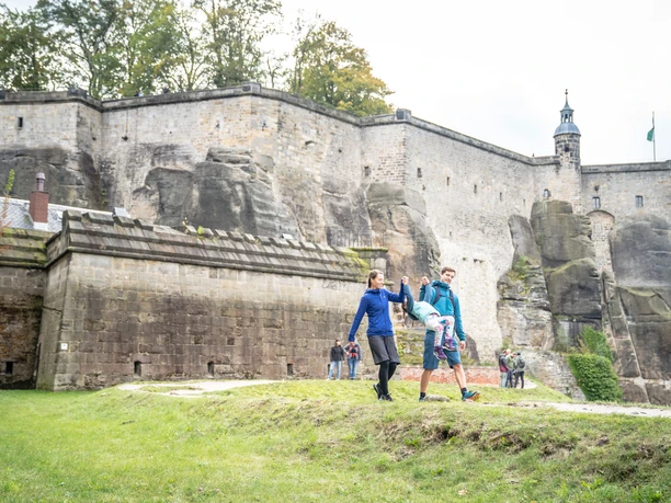 Rodina se prochází před historickým kamenným hradem na pevnosti Königstein, rodiče spokojeně houpají své dítě. Atmosféra je živá a uvolněná.