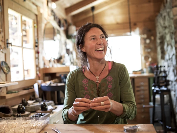 Susi´s Schmuckwerkstatt In einer Werkstatt sitzt eine lächelnde Frau beim Schmuckherstellen.