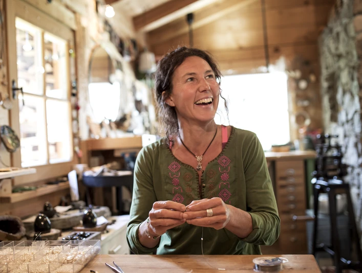 Susi´s Schmuckwerkstatt In einer Werkstatt sitzt eine lächelnde Frau beim Schmuckherstellen.