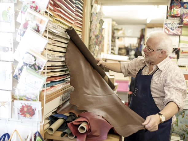 Buchbinderei Schreibwaren Matheisl Inser Hoamat Partner Ein älterer Mann in einer Buchbinderei betrachtet lederne Materialien.