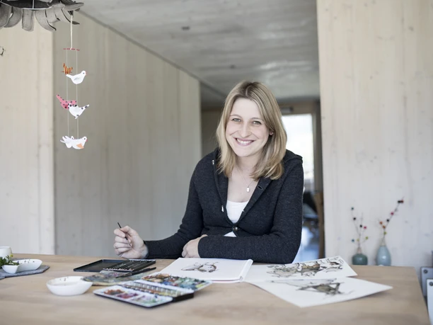 Karlmeise Inser Hoamat Partner Eine lachende Frau sitzt an einem Tisch mit Farben und Skizzen.