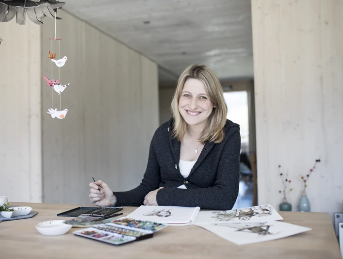 Karlmeise Inser Hoamat Partner Eine lachende Frau sitzt an einem Tisch mit Farben und Skizzen.