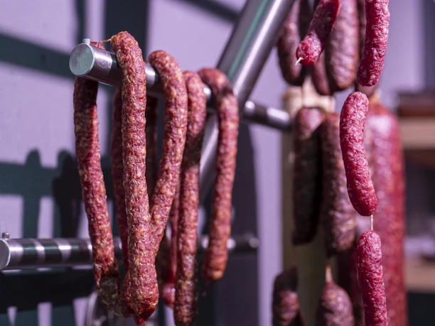 Hofladen beim Schweb Inser Hoamat Partner Geräucherte Würste hängen an metallischen Stangen in einem Hofladen.