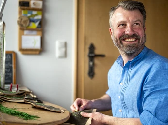 Lederhosen Michael Krippel Ein Mann in blauer Kleidung lächelt, während er Lederhosen an einem Tisch näht.