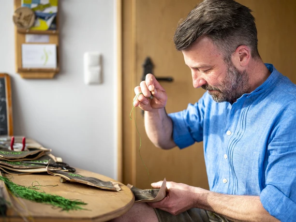 Lederhosen Michael Krippel Mann in blauer Kleidung näht Leder mit grünem Faden an einem Tisch.