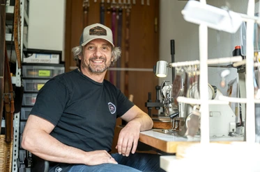 Handwerk & Design Bauer Ein Mann mit Baseballkappe sitzt lächelnd an einem Arbeitstisch mit Nähmaschine.