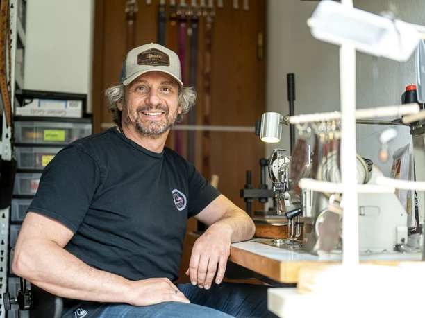 Handwerk & Design Bauer Ein Mann mit Baseballkappe sitzt lächelnd an einem Arbeitstisch mit Nähmaschine.
