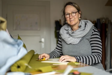 Stoff und Knopf Sabine Ritter Person in Werkstatt arbeitet an grünem Stoff, trägt Brille und gestreiftes Shirt.