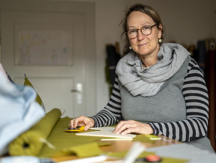 Stoff und Knopf Sabine Ritter Person in Werkstatt arbeitet an grünem Stoff, trägt Brille und gestreiftes Shirt.