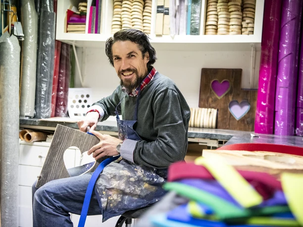 Alpenterieur Werkstatt mit Mann, der Holz bearbeitet, umgeben von Stoffen und Materialien.