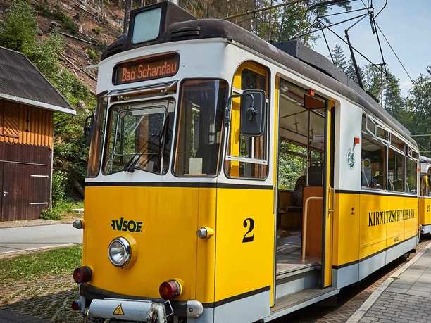 Yellow Kirnitzschtalbahn with the inscription "Bad Schandau" and "KIRNITZSCHTALBAHN" stands at a stop, surrounded by trees and a wooden building.