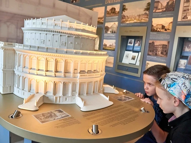 Dva chlapci si prohlížejí osvětlený model historické budovy v muzeu, obklopený informačními tabulemi a obrázky.