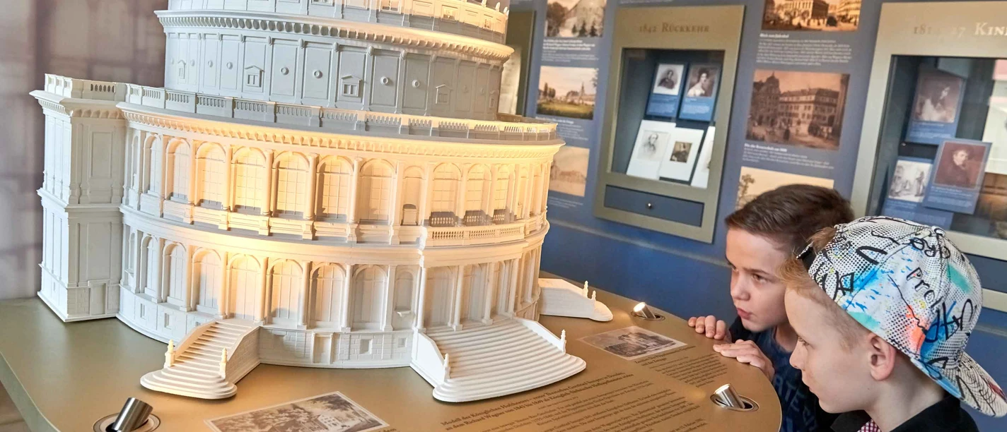 Modell der Semperoper Zwei Jungen betrachten ein beleuchtetes Modell eines historischen Gebäudes in einem Museum, umgeben von Informationstafeln und Bildern.