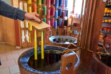 A hand dips two yellow candles in liquid wax; in the background, colorful candles are displayed on the wall.
