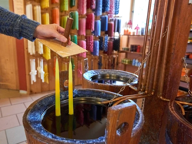 Kerzen selber ziehen Eine Hand taucht zwei gelbe Kerzen in flüssiges Wachs; im Hintergrund sind bunte Kerzen an der Wand ausgestellt.