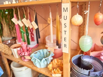 verschiedene Kerzen in der Kerzenzieherei Krietzschwitz Regal mit bunten Kerzen und dekorativen Wachsschalen, Schild mit "Muster" beschriftet, warme und einladende Atmosphäre.