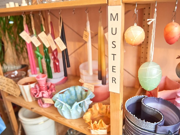 verschiedene Kerzen in der Kerzenzieherei Krietzschwitz Regal mit bunten Kerzen und dekorativen Wachsschalen, Schild mit "Muster" beschriftet, warme und einladende Atmosphäre.