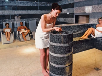 Sauna im Geibeltbad Pirna Sauna im Geibeltbad Pirna: Eine Frau im weißen Handtuch wäscht ihre Hände an einem grauen Brunnen in einem Spa, während zwei Personen im Hintergrund auf Liegen entspannen.
