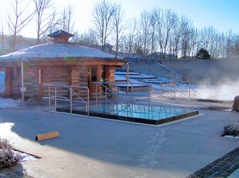 Geibeltbad Saunagarten im Winter Geibeltbad Pirna: Holzhütte neben einem dampfenden Außenpool in winterlicher Umgebung, umgeben von kahlen Bäumen und leichtem Schneefall.