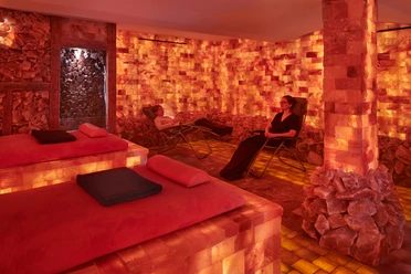 Two people relax on loungers in a warmly lit salt room with orange-colored walls and floors made of salt stones.