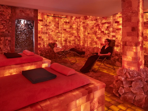 Salzscheune Berggießhübel Zwei Personen entspannen auf Liegestühlen in einem warm beleuchteten Salzraum mit orangefarbenen Wänden und Böden aus Salzsteinen.