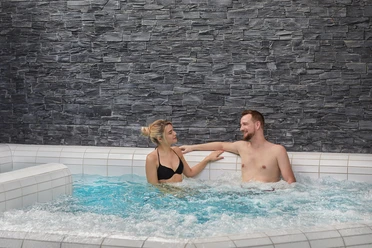 Een man en een vrouw ontspannen glimlachend in een bubbelbad voor een grijze stenen muur.